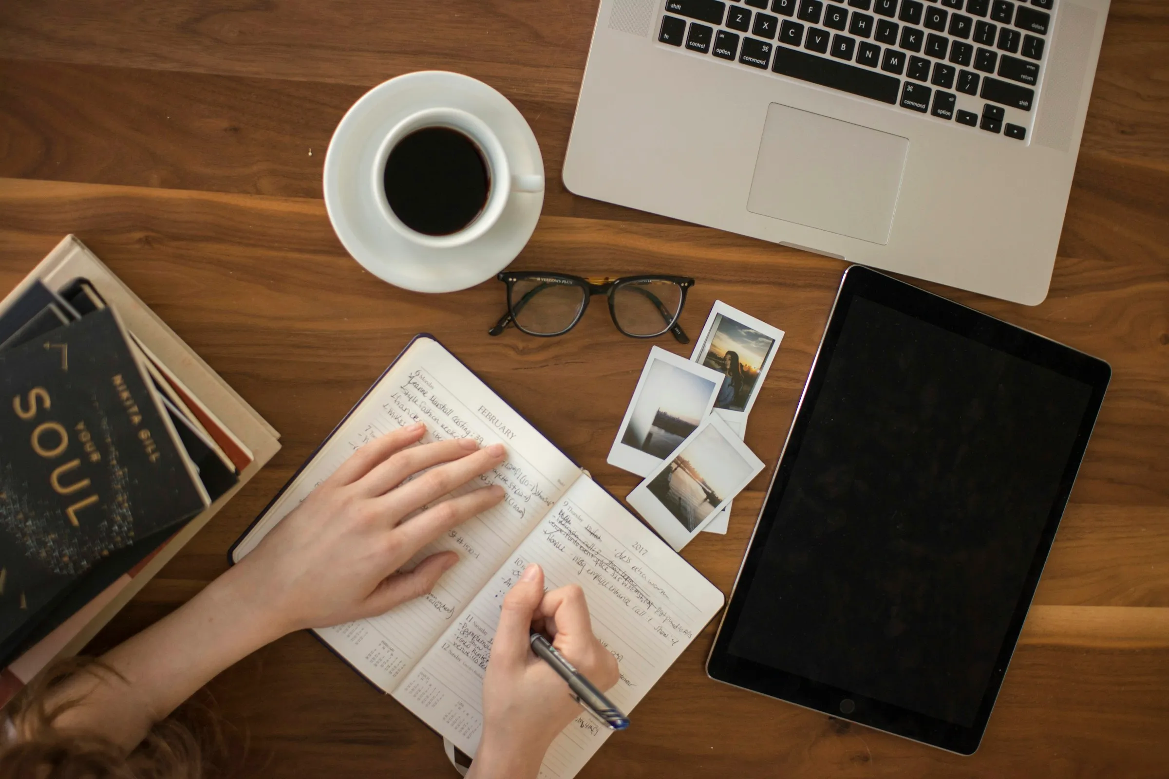 Hands writing in a notebook beside a laptop, tablet and coffee